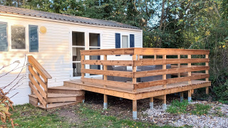Mobilheim Grand Confort mit Holzterrasse und Treppe, umgeben von Bäumen und Grün in der Natur.