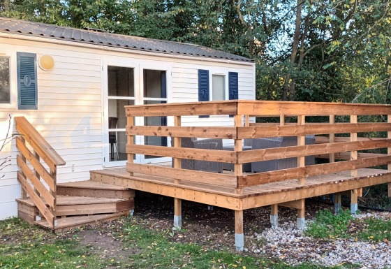 Mobil-home Grand Confort avec terrasse et escaliers en bois, entouré d'arbres et de verdure naturelle.