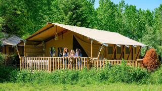 Drie mensen staan op de veranda van een grote safaritent in een groene, bosrijke omgeving in de natuur.