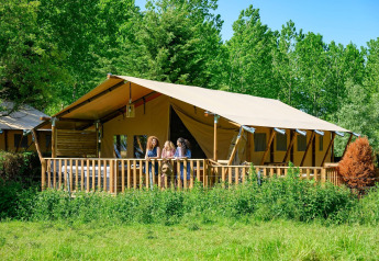Drie personen staan op het terras van een grote safaritent te midden van groene natuur en bomen buiten.