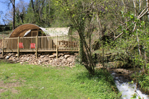 Tenda safari CocoSweet con terrazza in legno e sedie rosse, immersa nel bosco vicino a un ruscello.