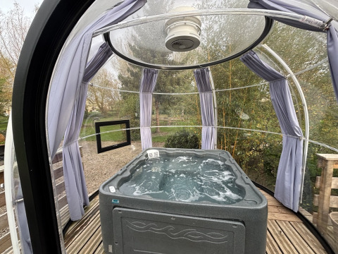 Bubbelbad in een transparante glampingkoepel met houten vloer, gordijnen en zicht op de tuin.