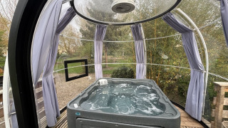 Bubbelbad in een transparante glampingkoepel met houten vloer, gordijnen en zicht op de tuin.