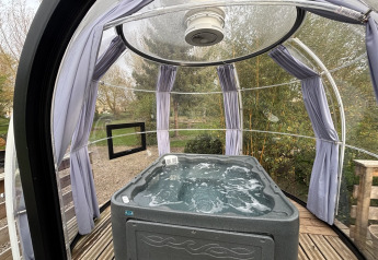 Bain à remous dans un dôme glamping transparent avec sol en bois, rideaux et vue sur le jardin.