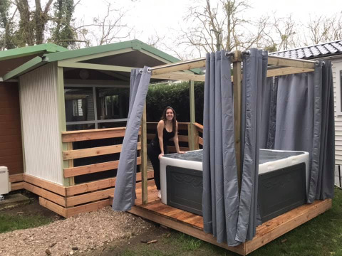 Vrouw bij een buitenjacuzzi met gordijnen bij Chalet du Bélier. Houten terras en groene omgeving.