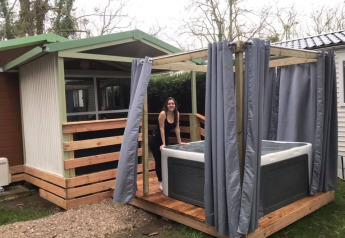 Kvinde ved udendørs jacuzzi med gardiner ved Chalet du Bélier. Træterrasse og grønne omgivelser.