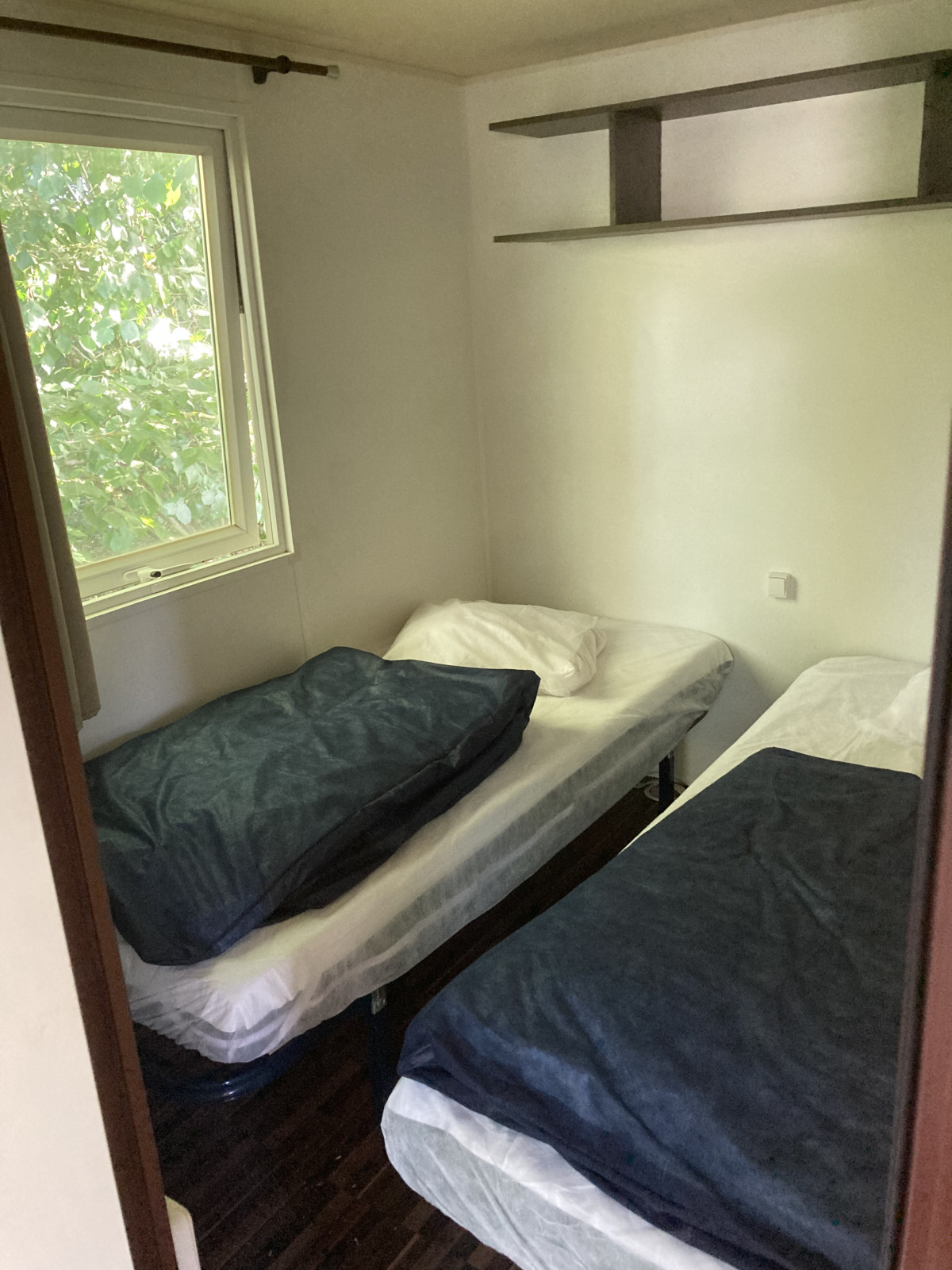 Kleine kamer met twee eenpersoonsbedden, raam met uitzicht op bomen en wandplank in een mobiel huis in Frankrijk.