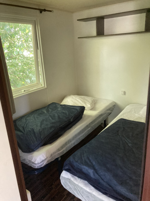 Small room with two single beds, a window facing trees, and a wall shelf in a mobile home in France.