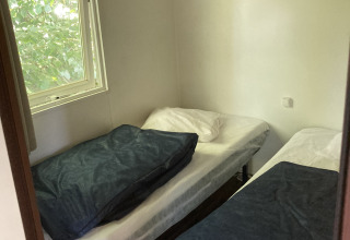 Petite chambre avec deux lits simples, fenêtre donnant sur des arbres et étagère murale dans un mobil-home.