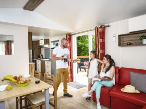 Gezin geniet in de living van Bungalow du Gorguet met moderne keuken, rode zetel en toegang tot groen terras.