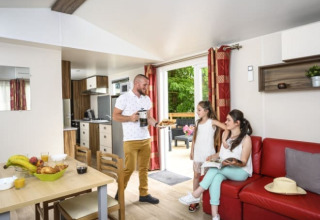 Gezin geniet in de living van Bungalow du Gorguet met moderne keuken, rode zetel en toegang tot groen terras.