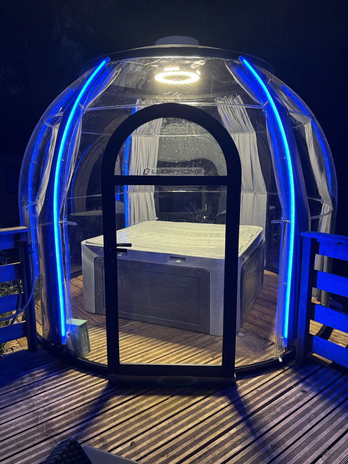 Transparante koepel met bubbelbad, verlicht door blauwe LED-verlichting bij nachtelijk glampingverblijf.
