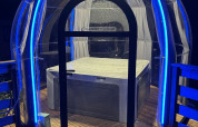 Transparante koepel met bubbelbad, verlicht door blauwe LED-verlichting bij nachtelijk glampingverblijf.