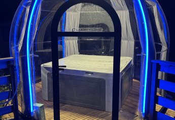 Dôme transparent avec jacuzzi à l'intérieur, éclairé par des LED bleues lors d'un séjour glamping de nuit.