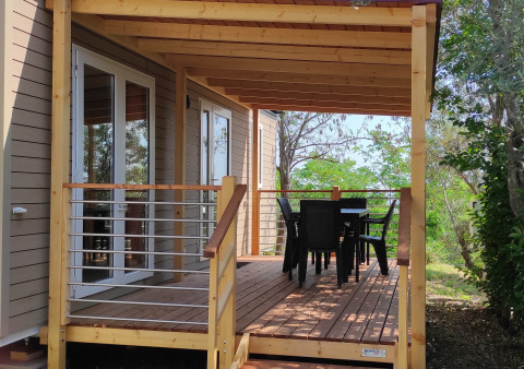 Casa mobile Riviera con veranda in legno, tavolo e sedie da esterno, circondata da alberi e verde.
