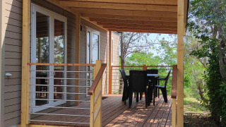 Mobil-home Riviera avec véranda en bois, table et chaises de terrasse, entouré d’arbres et de verdure.