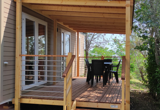 Mobile Home Riviera mit Holzveranda, Terrassentisch und Stühlen, umgeben von Bäumen und Natur.