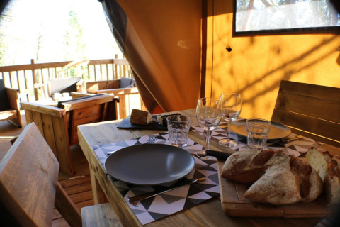 Gedekte tafel in safaritent met houten meubilair, glazen, borden en vers brood in zonlicht.