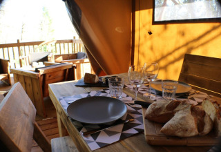 Set table in a safari tent with wooden furniture, glasses, plates, and fresh bread in sunlight.