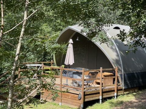 Tenda safari chiamata Lodge Big Oak XXL, circondata da alberi, con terrazza in legno e sedie da esterno.