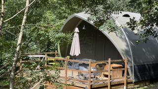 Tenda safari chiamata Lodge Big Oak XXL, circondata da alberi, con terrazza in legno e sedie da esterno.