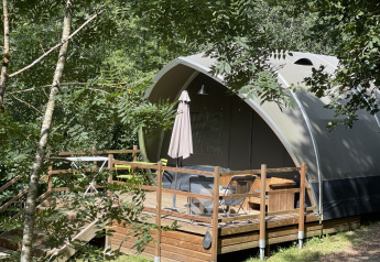 Tenda safari chiamata Lodge Big Oak XXL, circondata da alberi, con terrazza in legno e sedie da esterno.