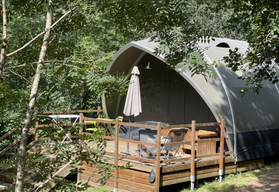 Tenda safari chiamata Lodge Big Oak XXL, circondata da alberi, con terrazza in legno e sedie da esterno.