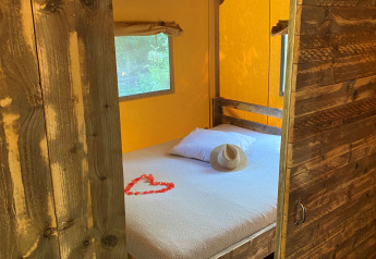 Interieur van Woody Safari Tent met wit bed, houten meubels, hoed en hart van rode bloemblaadjes.