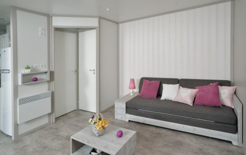 Lys stue i træhytte på Campsite Moulin de Chaules, med grå sofa, pink puder og minimalistisk indretning.