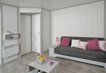 Bright living room in a wooden chalet at Campsite Moulin de Chaules, France, with grey sofa and pink cushions.