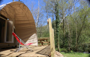 Terrazza soleggiata con sedia a sdraio rossa davanti a tenda CocoSweet al campeggio Moulin de Chaules, Francia.