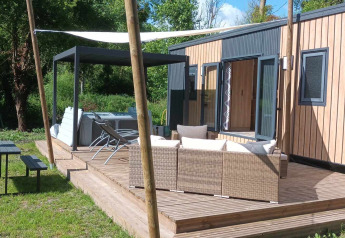 Sunny deck at Luxe Lodge with outdoor sofa, hot tub, dining table, and lush green surroundings.