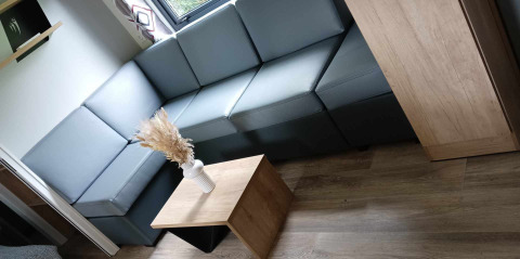 Moderne lodge-living met grijze hoekzetel, houten bijzettafeltje en vaas met droogbloemen aan het raam.