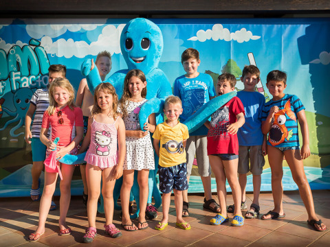 Een groep kinderen glimlacht met een grote blauwe octopusmascotte voor een kleurrijk buitenachtergrond.
