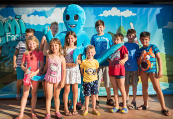 Een groep kinderen poseert met een groot blauw octopusmascotte voor een kleurrijke achtergrond.
