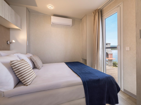 Moderne cabin met tweepersoonsbed, gestreepte kussens, blauw deken en uitzicht op het meer via de deur.