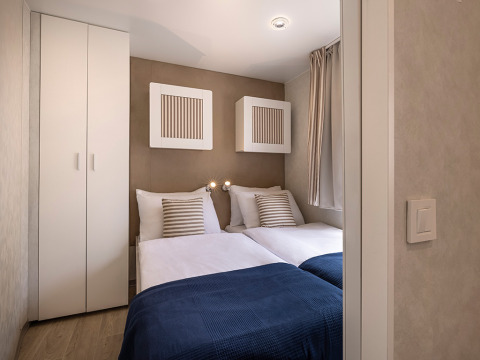 Kleine slaapkamer in een hut met twee eenpersoonsbedden, witte kast, blauwe dekens en gestreepte kussens.