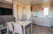 Zona pranzo e cucina moderna con quattro sedie, bottiglia di vino e TV in un lodge Park Riviera al campeggio Park Umag, Croazia.