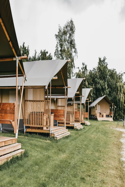Rangée de tentes safari avec toilettes sur un terrain de camping verdoyant, arbres et ciel nuageux.