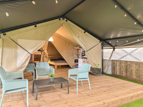 Tenda safari con lati aperti, area salotto moderna su pedana di legno e vista su letto e cucina interna.