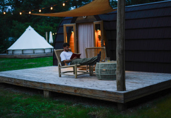 Hombre relajándose y leyendo en la terraza de una cabaña en Thuysgrond, Países Bajos, con luces nocturnas.