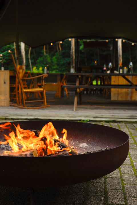 Feuerschale im Freien vor einem stilvollen Glamping-Bereich mit rustikalen Holzmöbeln und Naturblick.