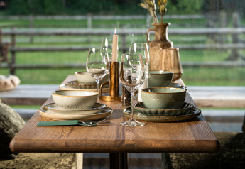 Elegant udendørs borddækning på glamping med keramiske tallerkener, skåle og glas mod grøn baggrund.