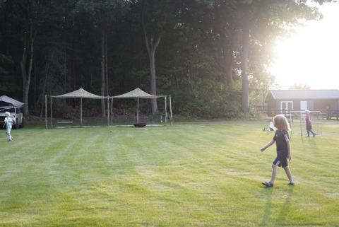 Kinderen spelen op het gras bij een glamping, met tenten en bosrand in de ondergaande zon.