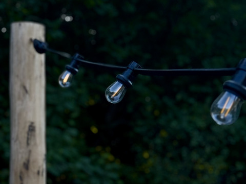 Guirnalda de luces exteriores en un poste de madera junto a una carpa Bell, rodeada de verde al atardecer.