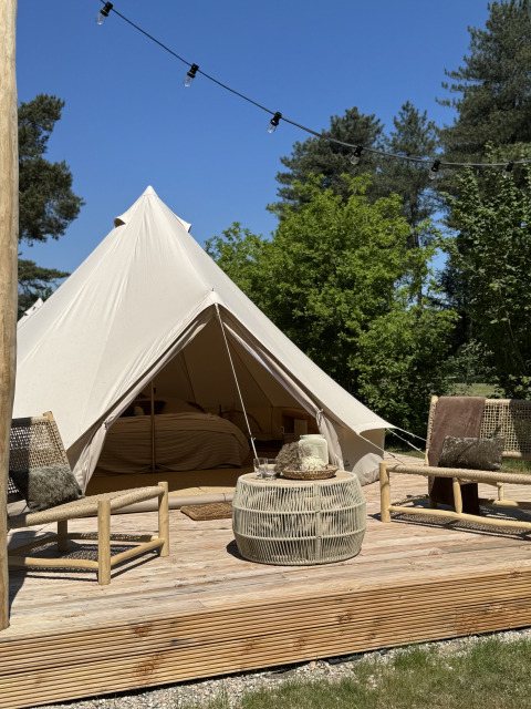 Ein Bell-Zelt-Tipi mit einer Holzterrasse, zwei Stühlen und einem Tisch, von Bäumen und blauem Himmel umgeben.