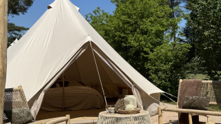 Tipi Bell tent con ingresso aperto, terrazza in legno, sedie e tavolino, circondato da alberi e cielo azzurro.