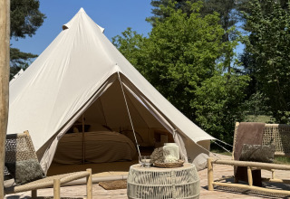 Tipi Bell tent avec entrée ouverte, terrasse en bois, chaises et table, entouré d’arbres sous ciel bleu.