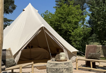 Ein Bell-Zelt-Tipi mit einer Holzterrasse, zwei Stühlen und einem Tisch, von Bäumen und blauem Himmel umgeben.