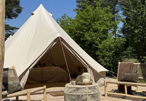 Bell tent tipi met open ingang, houten dek, stoelen en tafel, omringd door bomen en blauwe lucht.